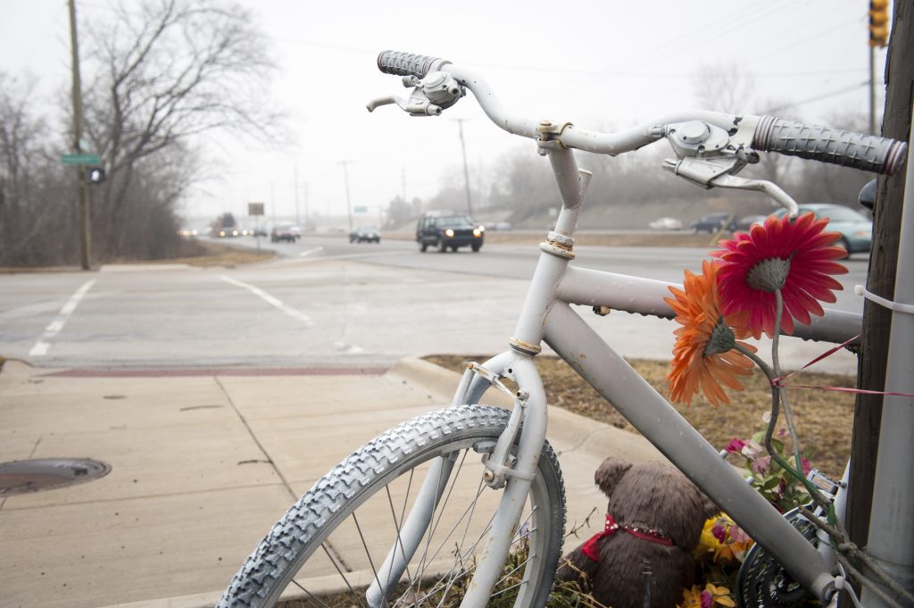 A white bike memorial leans on a post on a sidewalk near a busy road.